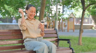 Park bankında telefon kullanırken Genç Kadın Heyecanını İfade Ediyor