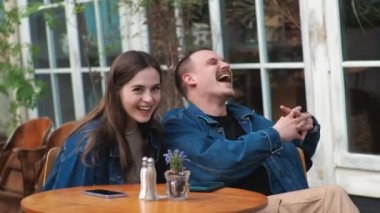 Genç bir çift. Denim giysileri. Eski bir restoranda randevuda, dışarıda. Konuşuyorlar. Sallanıyorlar.