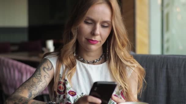 カジュアルな服装の若い女性がカフェの窓側に座ってコーヒーを飲んでいます 電話を見て ソーシャルネットワークにテキストを入力する — ストック動画