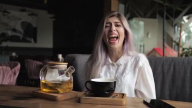 Genç sarışın iş kadını bir restoranda oturur ve pencerenin kenarında çay içer. Şaşırmış, gülümsüyor, dikkatsizce gülüyor, ağzı açık.