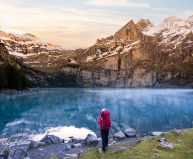 Sırt çantalı bir yürüyüşçü kız. Gün doğumunda İsviçre 'de dağ gölü yakınlarında. Arka planda bulutlu dağ zirveleri var. İsviçre Alplerinde Oeschinensee Gölü