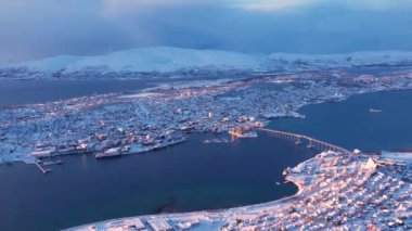 Norveç 'in Tromso kasabasının fiyort ve köprüsündeki karlı evlerin hava günbatımı manzarası. Güneşli kış havası