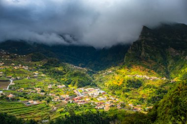 Geleneksel teras köyü Sao Vicente, Madeira Adası, Portekiz. Fırtınalı havada yeşil bir dağ manzarası arasında küçük evler ve bahçeler.