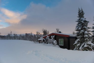 A small snow covered hunting lodge in the norwegian mountains with animal traces in the foreground and mountain summits in the background clipart