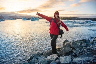 Kırmızı ceketli kadın sevinçle İzlanda 'daki bir buzul gölünün kayalık sahilinde poz veriyor. Geniş bir gülümseme içinde kollarını açıp gün batımının parıldayan güzelliğini yakalıyor.