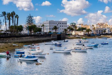Lanzarote 'deki Arrecife Lagoon' un canlı bir sahnesinde sayısız tekne berrak sularda huzur içinde yüzüyor. Modern binalar ve uzak dağlar sakin arkaplanı tamamlıyor