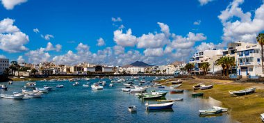 Lanzarote 'deki Arrecife Lagoon' un canlı bir sahnesinde sayısız tekne berrak sularda huzur içinde yüzüyor. Modern binalar ve uzak dağlar sakin arkaplanı tamamlıyor