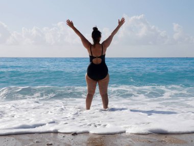 Siyah mayo giymiş kıvrımlı bir kadın denizde duruyor ve ellerini ufkun arka planına doğru kaldırıyor.