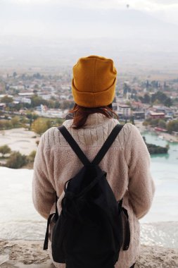 a girl with a black backpack in a yellow hat stands on top of a mountain in Pamukkale, rear view