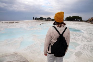 a girl with a black backpack in a yellow hat stands on top of a mountain in Pamukkale, rear view