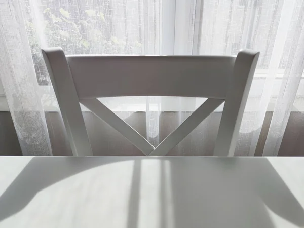 white chair at a white table against the background of a white translucent curtain in the sunlight.