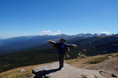 Neşeli, gülümseyen, yaşlı, elleri havada bir kadın dağın tepesinde duruyor..
