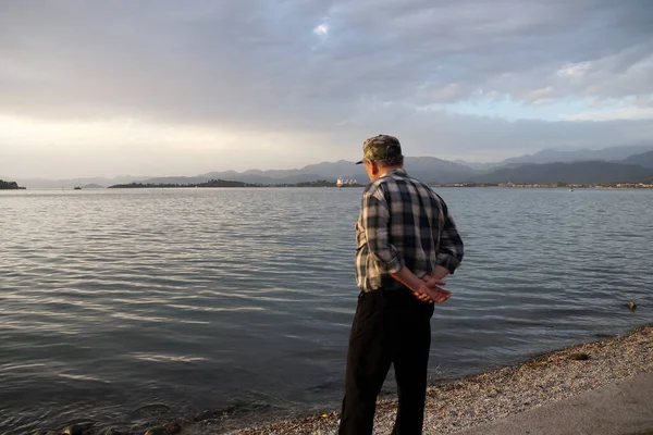 an elderly man stands on the seashore in a checkered shirt, rear view.