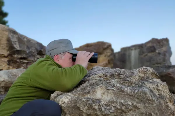 Büyük bir taşa yaslanan bir adam dürbünle bakar..