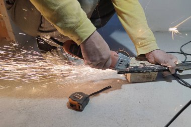 Bir adam metal bir profili öğütücüyle kesiyor, yakınlarda bir mezura var. Yakın çekim eller ve uçan kıvılcımlar.