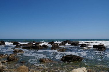 Dalgalar açık deniz ufkunun arka planına karşı kıyıdaki büyük kayalara çarpar..