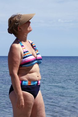 Çizgili mayo ve güneşlik takmış bir kadın deniz ufkunun önünde dikilirken güneşleniyor..