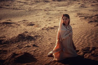 Dalgalı krem kumaşlı düşünceli kadın altın saatinde çöl kumunda oturur. Ilık ışık yüzünü ve çevresini aydınlatıyor. Yalnızlığı, içselliği ve doğayla bağlantıyı taşır..