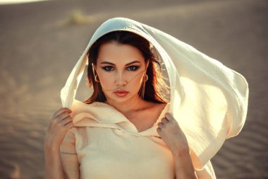 Çöl manzaralı güzel bir kadın, açık renk kumaş elbise ve duvak giyiyor. Yoğun bakışlar, güneş yanığı tenler. Göksel ve gizemli bir atmosfer. Moda, güzellik, seyahat ve yaşam tarzı temaları için mükemmel. Altın ışıkta egzotik ve çekici bir portre..