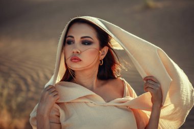 Çöl manzaralı güzel bir kadın, açık renk kumaş elbise ve duvak giyiyor. Yoğun bakışlar, güneş yanığı tenler. Göksel ve gizemli bir atmosfer. Moda, güzellik, seyahat ve yaşam tarzı temaları için mükemmel. Altın ışıkta egzotik ve çekici bir portre..