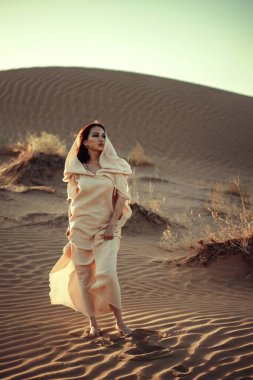 Akan beyaz elbiseli kadın altın saatinde çölde yürüyor. Yumuşak ışık, yüz hatlarını ve kumaş dokusunu geliştirir. Zarafet, gizem ve macera taşır. Moda ve seyahat temaları için ideal.