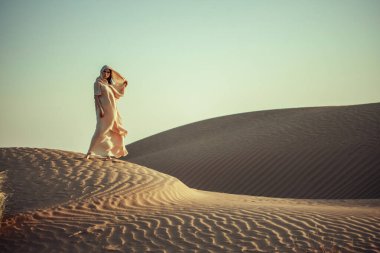 Çöl tepesinde uçan elbiseli bir kadın. Altın ışık, dalgalı kum. Macera, yalnızlık ve ebedi güzelliği çağrıştırıyor. Seyahat ve yaşam tarzı temaları için mükemmel.