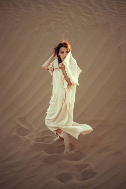 Çöl zeminine karşı beyaz kumaşlı ilahi bir kadın. Yoğun bakışlar, zarif duruş. Gizem ve zarafeti harmanlıyor. Moda, kültür ve sanatsal temalar için mükemmel. Çöl güzelliğinin cazibesini yakalar..