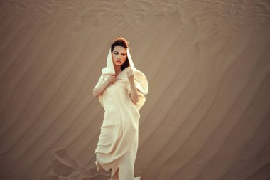 Çöl zeminine karşı beyaz kumaşlı ilahi bir kadın. Yoğun bakışlar, zarif duruş. Gizem ve zarafeti harmanlıyor. Moda, kültür ve sanatsal temalar için mükemmel. Çöl güzelliğinin cazibesini yakalar..