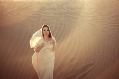 Akan beyaz kumaşlı ruhani kadın altın saatinde çöl kumullarının arasında duruyor. Gizemli ve cezbedici bakışları ufukta belirdi. Yumuşak ışık sahneyi yıkıyor, rüya gibi bir atmosfer yaratıyor. Seyahat, moda ve ruhani temalar için mükemmel..