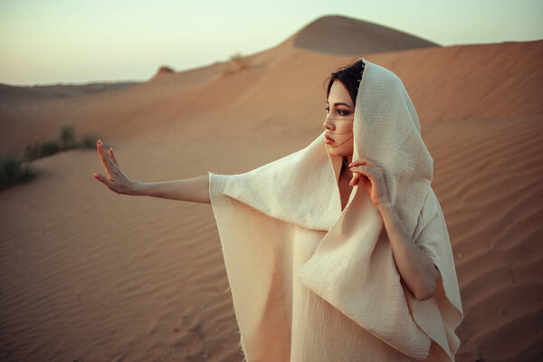 Beautiful woman in desert landscape, wearing light fabric dress and veil. Intense gaze, sun-kissed skin. Ethereal and mysterious atmosphere. Perfect for fashion, beauty, travel, and lifestyle themes. Exotic and alluring portrait in golden light.
