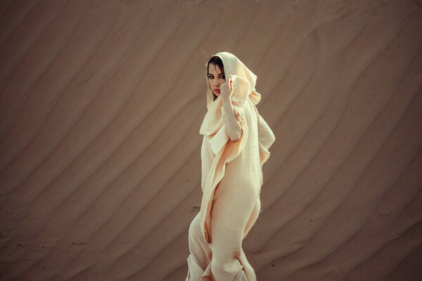 Ethereal woman in flowing white fabric against desert backdrop. Intense gaze, graceful pose. Blends mystery and elegance. Perfect for fashion, cultural, and artistic themes. Captures allure of desert beauty.