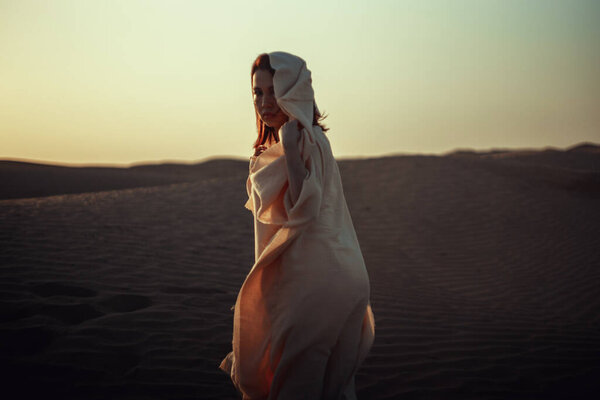 Ethereal woman in flowing white fabric against desert backdrop. Intense gaze, graceful pose. Blends mystery and elegance. Perfect for fashion, cultural, and artistic themes. Captures allure of desert beauty.