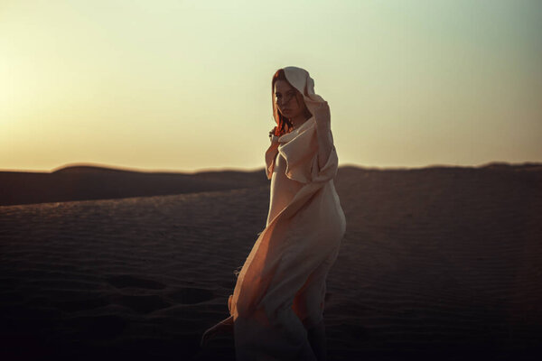Ethereal woman in flowing white fabric against desert backdrop. Intense gaze, graceful pose. Blends mystery and elegance. Perfect for fashion, cultural, and artistic themes. Captures allure of desert beauty.