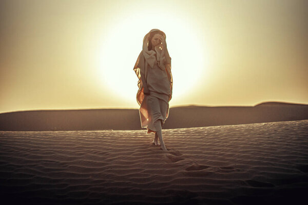 Woman in flowing dress silhouetted against radiant sunset in desert. Ethereal atmosphere as she lifts fabric. Embodies freedom, spirituality, and connection with nature. Ideal for lifestyle and travel themes.
