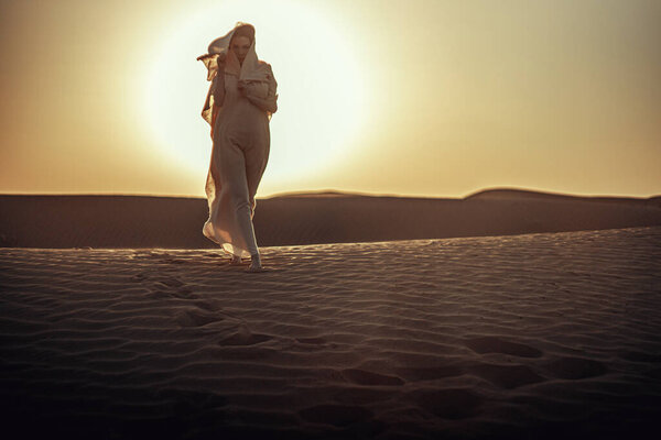 Woman in flowing dress silhouetted against radiant sunset in desert. Ethereal atmosphere as she lifts fabric. Embodies freedom, spirituality, and connection with nature. Ideal for lifestyle and travel themes.