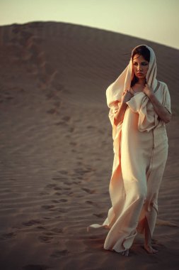 Çöl kumulunda yürüyen beyaz elbiseli bir kadın. Altın ışık ruh halini geliştirir. Kumda ayak izleri yolculuğun hikayesini anlatır. Yalnızlık, macera ve kültürel zenginlik taşır. Seyahat ve moda temaları için mükemmel.