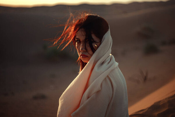 Intense close-up of woman in desert, hair tousled by wind. White garment and warm light create striking contrast. Her gaze is powerful and enigmatic. Ideal for portraying strength, beauty, and the untamed spirit of nature.