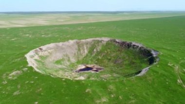 View of the funnel from a nuclear explosion, overgrown with green grass and a lake in the center of the funnel. There are many small craters around the large crater. large meteorite impact crater