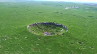 View of the funnel from a nuclear explosion, overgrown with green grass and a lake in the center of the funnel. There are many small craters around the large crater. large meteorite impact crater