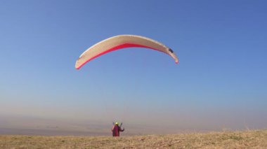 The pilot on the paraglider catches the tailwind, keeps the wing on top, trying to cherish.Above the paraglider there is a blue cloudless sky and autumn fields.