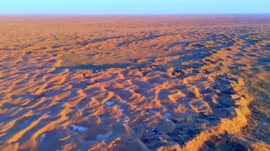 Gün batımında kum tepeleri olan bir çöl manzarası, altında araçlar olan bir kamp. Doğa, seyahat ve macera temaları için çarpıcı bir manzara.