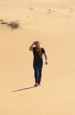 Şapkalı bir adam alevli güneşin altında çöl kumullarında yürür, şapkasını rüzgara karşı tutar.