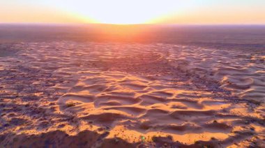 Gün batımında çöl kumullarının üzerinde insansız hava aracı, aşağıda arabalarla kamp. Vahşi doğanın, doğanın ve keşiflerin büyüleyici manzarası.