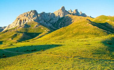 Gün batımında parlayan keskin dağ zirveleri, açık mavi gökyüzü ile kontrastlı yeşil yamaçlar