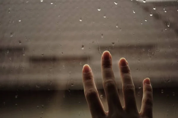 Hand touches a window with rain drops. Roof background in blur.