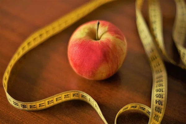 Red apple and yellow measuring tape on the brown wooden table.
