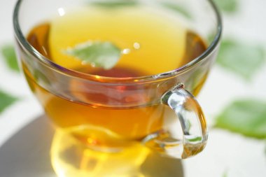 Huş yaprağıyla birlikte cam bardakta yeşil çay. Bardağın etrafında beyaz bir masada içkiyle duruyor..