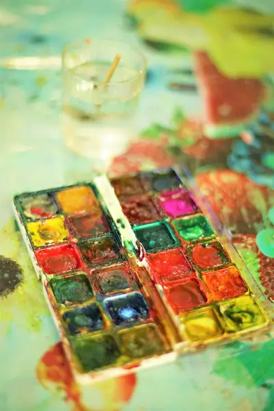 Old dirty watercolor paint set and glass of water with a brush on the table.