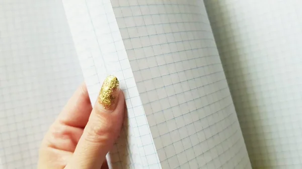 A beautiful female hand with a golden manicure flips through a blank checkered notebook