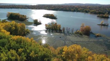 Mille Iles nehri üzerinde sağdan sola dönen hava aracı görüntüsü. Suda güneş yansıması ve sonbaharın renkli ağaçları var.